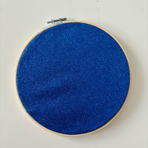 May include: A round wooden embroidery hoop with blue glitter fabric stretched across the inside.