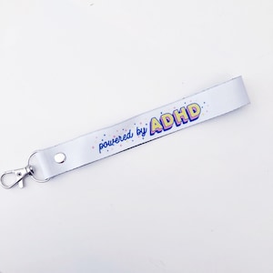 Può includere: Lacci bianco con un moschettone argentato. Il laccio presenta una grafica gialla, rosa e blu che dice "powered by ADHD" con piccole stelle rosa e gialle.