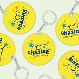 Può includere: Quattro portachiavi rotondi gialli con una catena argentata. Ogni portachiavi presenta un'illustrazione colorata di una molecola di dopamina con il testo "chasing dopamine" in un carattere blu arcobaleno.