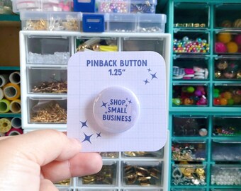 Könnte beinhalten: Ein 1,25 Zoll großer Ansteckknopf mit weißem Hintergrund und blauem Text, der "Shop Small Business" lautet. Der Knopf ist von kleinen blauen Sternen umgeben.
