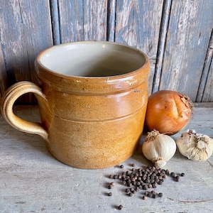 May include: A tan ceramic mug with a handle and horizontal grooves. An onion, two garlic bulbs, and peppercorns are arranged beside the mug. The mug has a light-coloured interior and sits on a wooden surface.