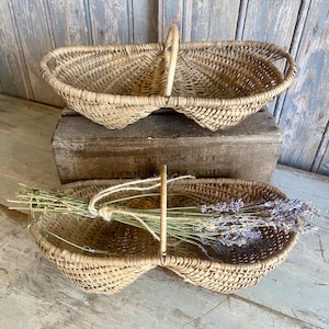 May include: Two oval-shaped woven baskets made of light brown natural reeds. Each basket features a central vertical handle and a divided interior structure. One basket contains a bundle of dried lavender stems tied with twine.