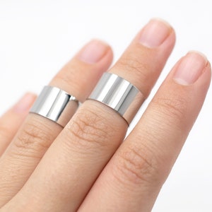 May include: Two silver-colored, wide band rings are shown on fingers against a white background. The rings have a smooth, polished surface and a minimalist design. The rings are made of a shiny metal.