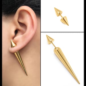 Puede incluir: Un par de pendientes de espiga de tono dorado. Los pendientes están hechos de metal y tienen un diseño puntiagudo y afilado. Los pendientes se muestran en la oreja de una modelo.