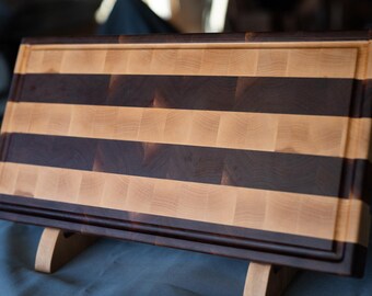 May include: A rectangular end-grain cutting board with alternating stripes of light and dark wood. The board has a recessed border and is displayed on a wooden stand. This butcher block style board is ideal for food preparation.