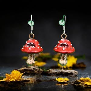 May include: A pair of mushroom-shaped earrings. The red caps have white spots, and the stems are detailed. The earrings hang from green and gold ear wires. The background is dark, with yellow flowers in the foreground.
