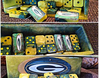 May include: A set of dominoes in a matching storage box, featuring a green and yellow marbled resin finish. Several dominoes display the Green Bay Packers logo, a white letter G inside a green oval with a yellow outline, on their faces.
