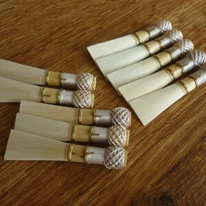 Puede incluir: Una colección de cañas de oboe de madera, dispuestas sobre una superficie de madera. Cada caña presenta una lámina rectangular de color claro, una banda dorada y un extremo decorativo tejido. Las cañas están agrupadas en dos conjuntos, mostrando su diseño y artesanía.