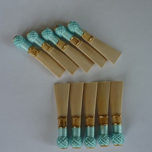 Puede incluir: Un conjunto de cañas de oboe. Las cañas son de color madera clara, con hilo turquesa enrollado alrededor de la base y una bola decorativa turquesa en el extremo. Están dispuestas sobre una superficie blanca.