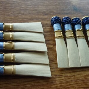 Puede incluir: Una colección de cañas de clarinete, con un color de madera natural y una punta rectangular clara. Cada caña tiene un extremo texturizado azul oscuro, detalles plateados y dorados, y está dispuesta sobre una superficie de madera.
