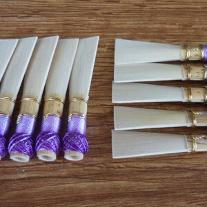 Puede incluir: Un conjunto de cañas de oboe, con una lámina rectangular de color claro unida a una base cilíndrica. La base está envuelta con hilo morado y asegurada con una banda dorada. Las cañas están dispuestas sobre una superficie de madera.