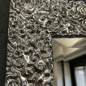 May include: Close-up of a silver mirror frame with a textured, swirling design. The frame has a reflective, metallic finish. The mirror's edge is visible, reflecting the surrounding environment. The frame's intricate pattern adds a decorative element.