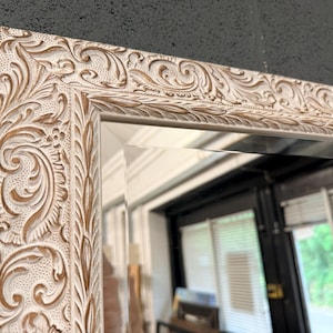 May include: A rectangular mirror with a detailed, carved frame. The frame has an ornate, floral design in cream with brown accents. The mirror reflects a room with windows and natural light.