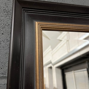 May include: A black framed mirror with a gold trim. The mirror has a bevelled edge and is reflecting a room with a white wall and a window.