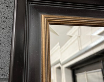 May include: A black framed mirror with a gold trim. The mirror has a beveled edge and is reflecting a room with a white wall and a window.