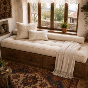 May include: A wooden daybed with built-in storage drawers, topped with a tufted cream cushion, a long bolster pillow, two square cream pillows, and a cream-colored knit throw blanket with fringe, positioned beneath a large window.