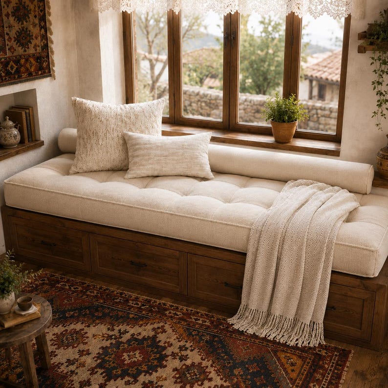 May include: A wooden daybed with built-in storage drawers, topped with a tufted cream cushion, a long bolster pillow, two square cream pillows, and a cream-colored knit throw blanket with fringe, positioned beneath a large window.