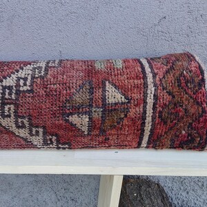 May include: A red, white, and brown patterned rug with a geometric design. The rug is folded in half and placed on a white wooden bench.