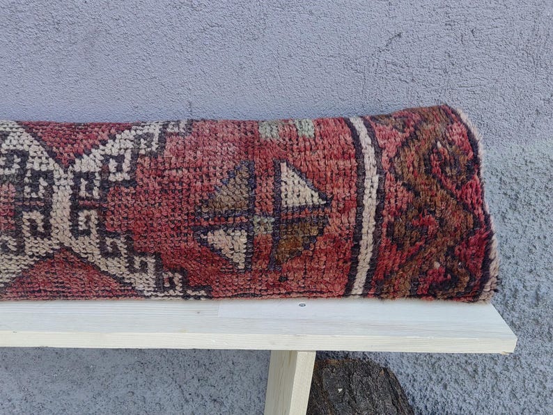 May include: A red, white, and brown patterned rug with a geometric design. The rug is folded in half and placed on a white wooden bench.