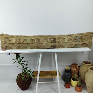 May include: A long, rectangular, beige and brown patterned cushion. The cushion is placed on a white table. The cushion has a repeating pattern of geometric shapes. The cushion is a decorative item.
