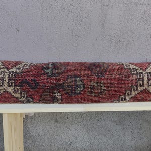 May include: A long, rectangular throw pillow with a red, beige, and brown patterned fabric. The pillow is sitting on a wooden bench.