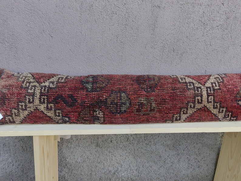 May include: A long, rectangular throw pillow with a red, beige, and brown patterned fabric. The pillow is sitting on a wooden bench.