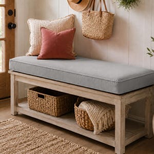 May include: A rectangular wooden entryway bench with a light gray upholstered seat cushion. The bench features a lower shelf containing two woven wicker storage baskets, one of which holds a folded cream-colored textile with fringe detailing.