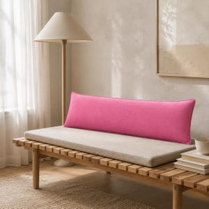 May include: A long, rectangular bolster cushion in a vibrant, solid hot pink colour. The cushion features a smooth, soft-touch fabric texture and is positioned horizontally across the length of a light beige bench cushion, providing a bold colour contrast.