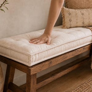 May include: A rectangular, off-white tufted bench cushion with visible stitching along the edges, placed on a dark brown wooden bench. The cushion features a soft, fabric texture with button-tufted detailing across its surface.
