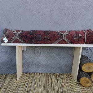 May include: A wooden bench with a red, black, and white patterned cushion. The bench is sitting on a woven bamboo mat.