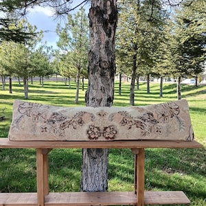Puede incluir: Un cojín rectangular beige y marrón con estampado floral descansa sobre una estantería de madera. El cojín presenta un diseño floral y un aspecto vintage. El fondo muestra un árbol y una zona de césped.