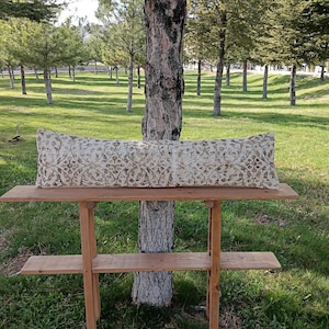 May include: A rectangular beige and brown patterned pillow rests on a two-tiered wooden table. The table is outdoors, with a tree trunk behind it and a grassy field with trees in the background.