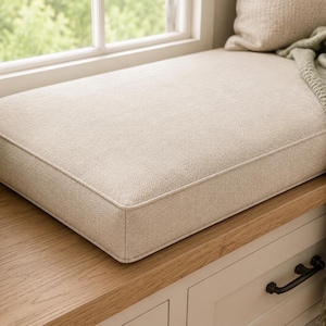 Puede incluir: Un cojín rectangular de tela beige con bordes ribeteados, colocado sobre un banco de madera. El cojín tiene un tejido texturizado y se acompaña de una almohada beige a juego y una manta de punto verde salvia con flecos.