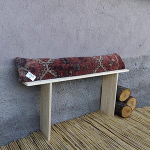 May include: A wooden bench with a red, brown, and white patterned cushion. The bench is made of light wood and has a simple design. The cushion is a traditional Turkish kilim rug.
