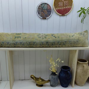 May include: A long, rectangular, light blue and yellow patterned bolster pillow sits on a wooden bench. The pillow is made of woven fabric and has a fringe on each end. The bench is made of light wood and has a simple design.