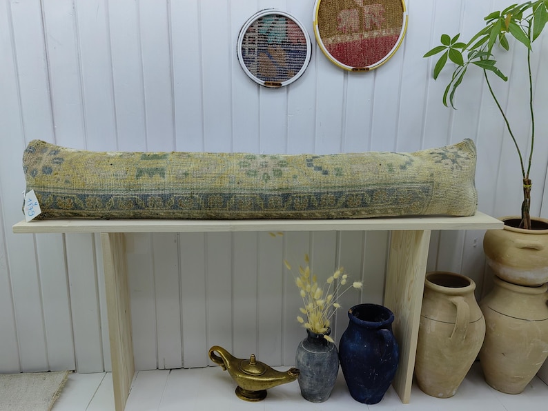 May include: A long, rectangular, light blue and yellow patterned bolster pillow sits on a wooden bench. The pillow is made of woven fabric and has a fringe on each end. The bench is made of light wood and has a simple design.