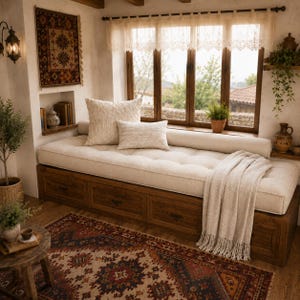 May include: A wooden daybed with three storage drawers underneath, topped with a tufted cream cushion, two rectangular throw pillows, and a cream-colored fringed throw blanket. The daybed is positioned against a window with lace curtains.