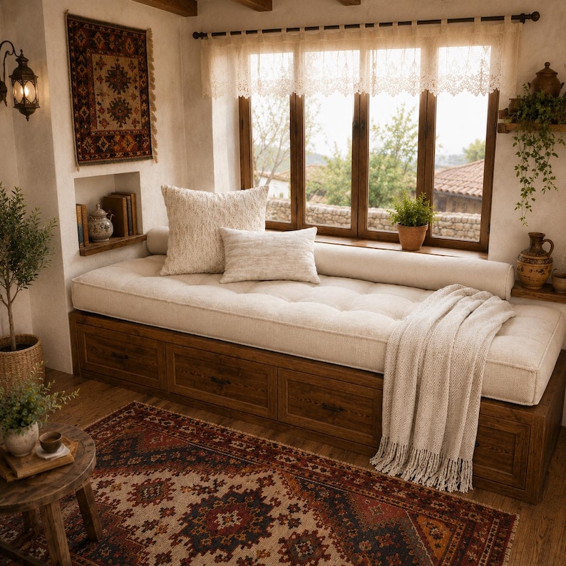 May include: A wooden daybed with three storage drawers underneath, topped with a tufted cream cushion, two rectangular throw pillows, and a cream-colored fringed throw blanket. The daybed is positioned against a window with lace curtains.
