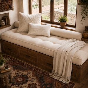 May include: A wooden daybed with three storage drawers underneath, featuring a tufted beige cushion, a large square textured pillow, a smaller rectangular beige pillow, and a beige herringbone throw blanket with fringe draped over the side.