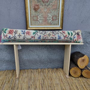 May include: A wooden bench with a floral patterned cushion. The bench is made of light wood and has a simple design. The cushion is a long, rectangular shape and is covered in a fabric with a floral pattern in shades of pink, green, and blue.