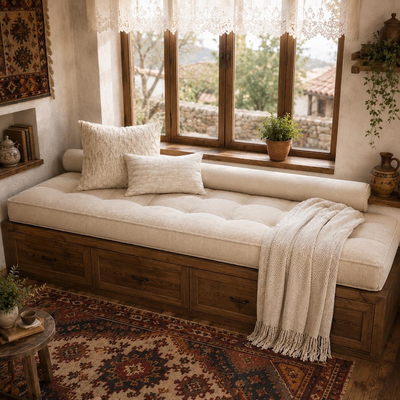 May include: A wooden window seat with three storage drawers underneath, topped with a tufted beige cushion, a long bolster pillow, two square throw pillows, and a cream-colored knit throw blanket with fringe, positioned in front of a window.