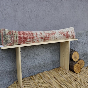May include: A long, rectangular cushion with a distressed red and beige pattern rests on a light wood table. The cushion's design has a vintage, rug-like appearance. The table is supported by two legs and a small stack of logs.