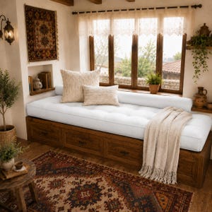May include: A wooden window seat with built-in storage drawers, topped with a long white tufted cushion. Two beige textured throw pillows rest on the cushion, accompanied by a cream-coloured fringed throw blanket draped over the edge.