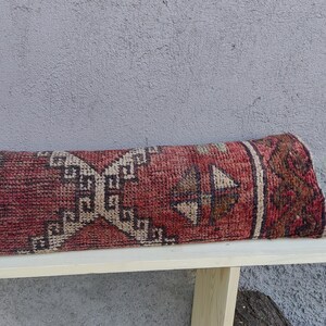 May include: A red, beige, and brown patterned rug rolled up on a wooden bench. The rug has a geometric design with a central diamond shape.