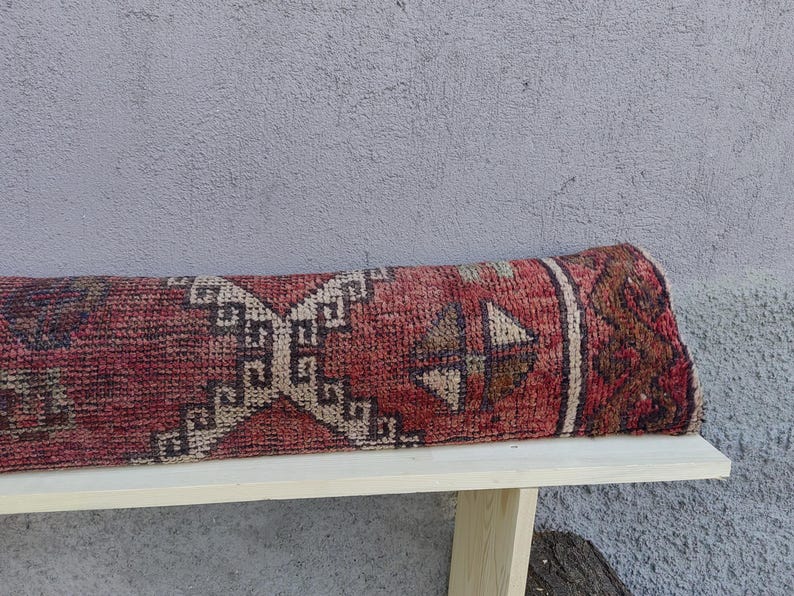 May include: A red, beige, and brown patterned rug rolled up on a wooden bench. The rug has a geometric design with a central diamond shape.