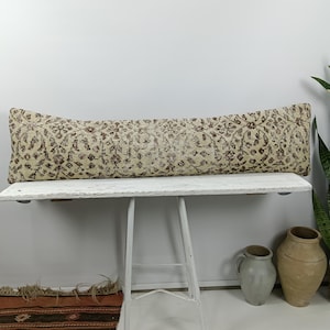 May include: A long, rectangular cushion with a beige background and a repeating brown floral pattern. The cushion is displayed on a white table. Decorative vases and a potted plant are in the background.