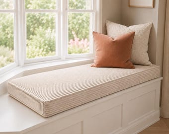 May include: A rectangular window seat cushion upholstered in a beige corduroy fabric. The cushion is placed on a white wooden bench and is topped with two square throw pillows, one in a solid terracotta orange and one in a textured cream fabric.