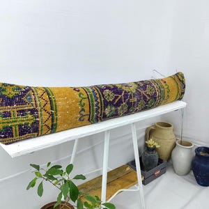 May include: A long, yellow and purple patterned bolster pillow sits on a white wooden bench. The pillow is made of woven fabric with a geometric design.