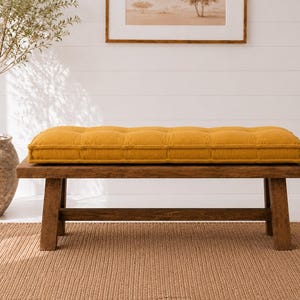 May include: A rectangular mustard yellow tufted bench cushion sits atop a dark brown wooden bench. The cushion features a grid-like pattern of tufted indentations across its surface, providing a thick, padded appearance for the wooden seat.