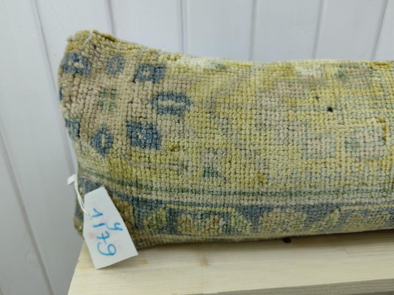 May include: A long, rectangular pillow with a blue and yellow patterned design. The pillow is made of a soft, woven fabric and has a tag attached to it that says "6+11".